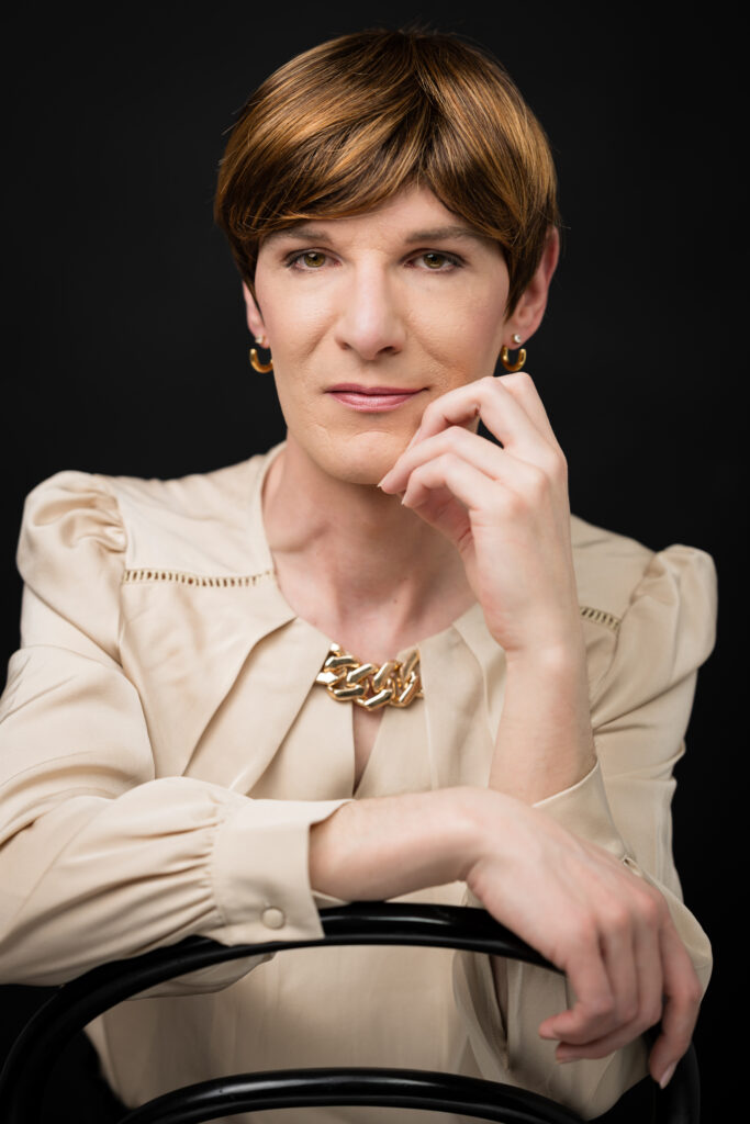 Porträt einer selbstbewussten Frau mit kurzem braunen Haar, beiger Bluse und goldenen Accessoires vor dunklem Hintergrund